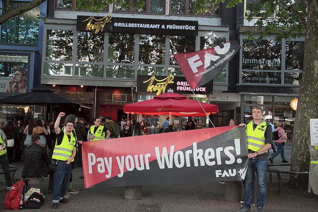Das Foto zeigt Teile der Kundgebung vor dem Keyf am 5. Juni mit einem Transparent, auf dem der Text "Pay your Workers! - FAU" zu lesen ist. Daneben verschiedene Aktivist*innen und Passanten.