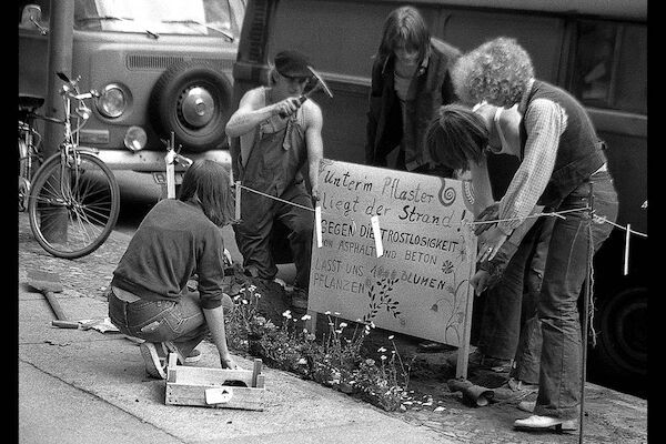 Ein paar Menschen stellen ein Plakat auf. Der Text: "Unter dem Pflaster liegt der Strand - gegen die Trostlosigkeit von Asphalt und Beton - lasst uns 1000 Blumen pflanzen".  Foto: © Monika von Wegerer  |  Schöneberg, Pallasstr., Aktion "unter dem Pflaster liegt der Strand," 1979