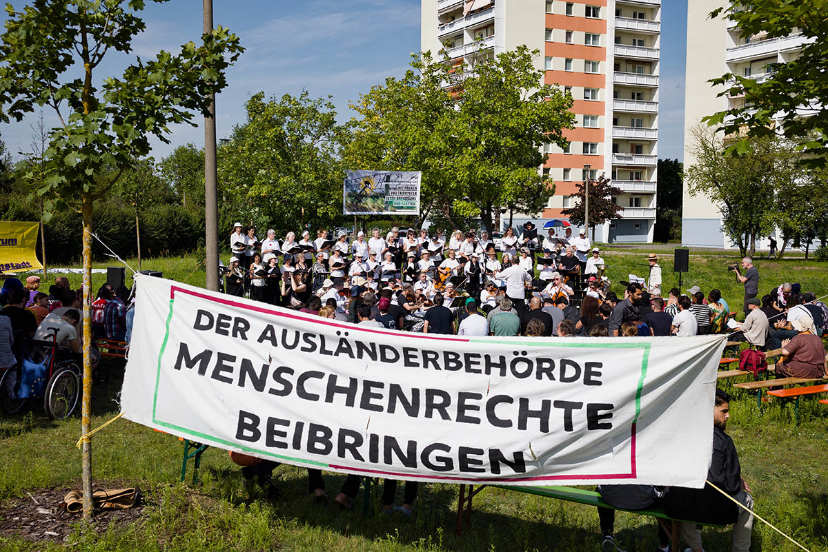 Das Foto zeigt das Konzert der Lebenslaute hinter dem Trasparent mit dem Text "Der Ausländerbehörde Menschenrechte beibringen". Im Hintergrund ist ein weiteres Transparent zu sehen sowie einige Häuser, darunter ein Hochhaus.