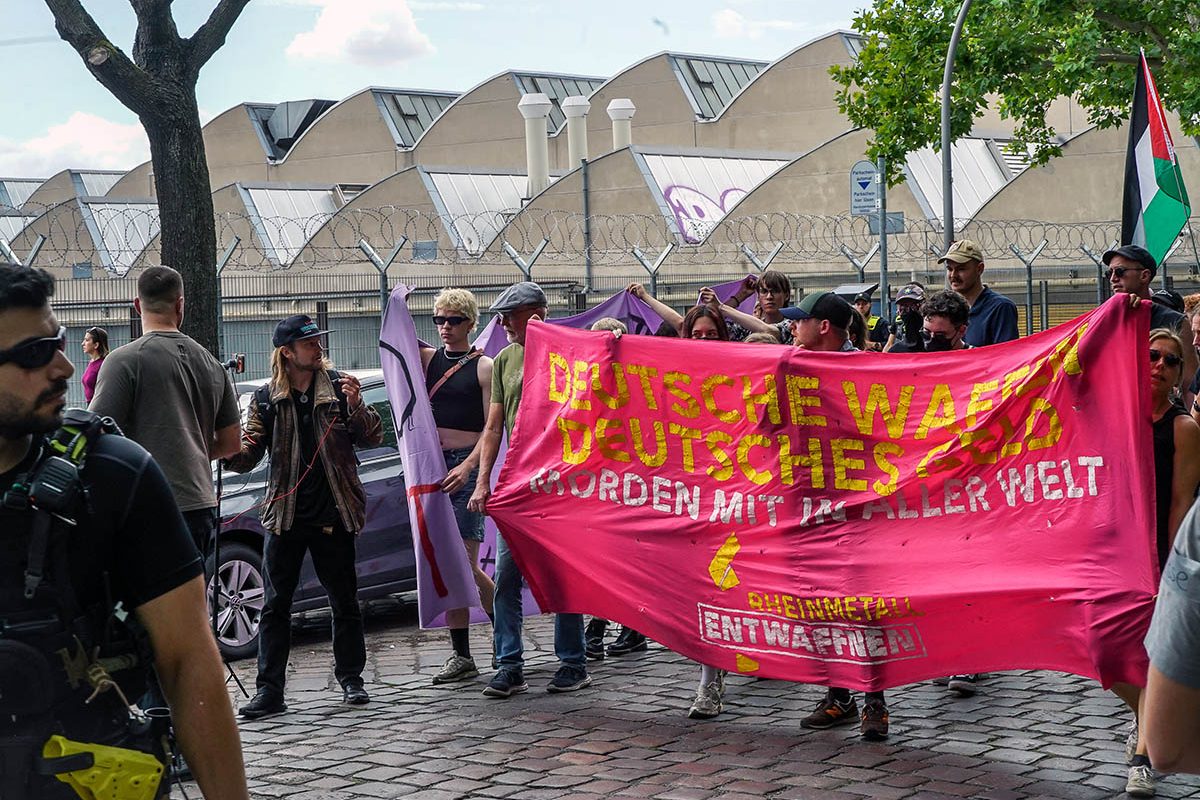 Das Foto von © heba zeigt eine Demonstration vor Fabrikhallen mit einem Fronttransparent auf dem der Text "Deutsche Waffen deutsches Geld morden mit in aller Welt" sowie das Logo von "Rheinmetall entwaffnen" zu lesen ist. links im Vordergrund ein Polizist mit Sonnenbrille, der Richtung Demo schaut, hinter dem Transparent sind einige Menschen sowie eine Palästina Fahne zu sehen.