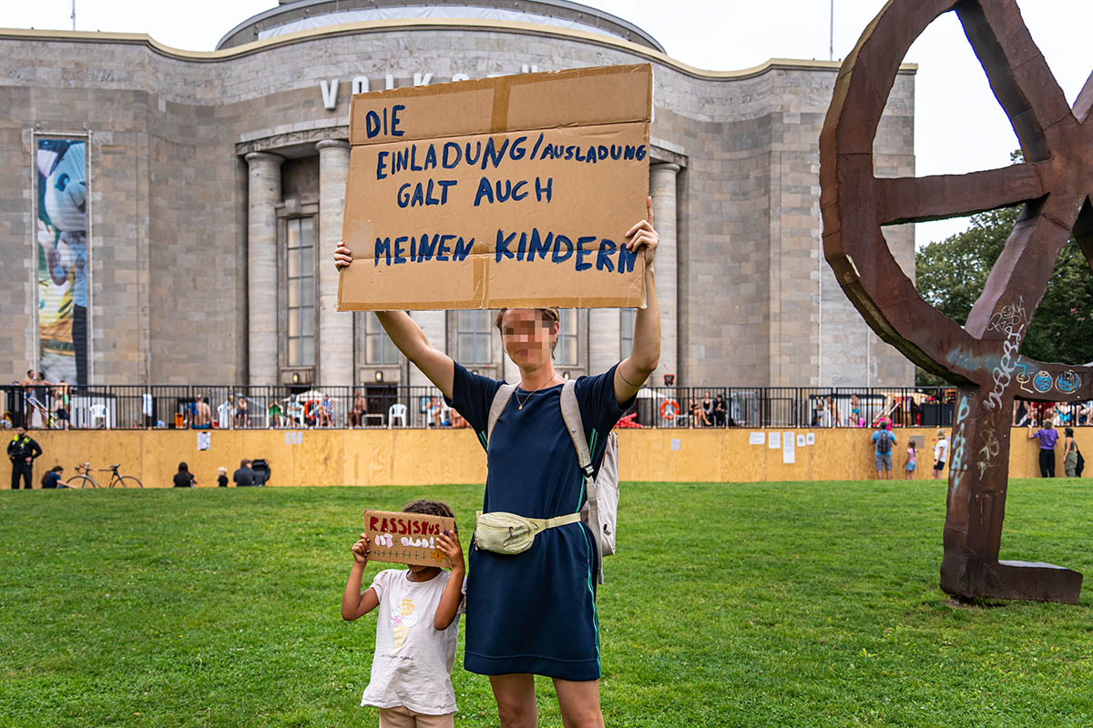 Das Foto von © Björn Obmann zeigt eine Frau mit Kind. Die verpixelte Frau hält eine Papptafel mit dem Text "Die Einladung / Ausladung galt auch meinen Kindern" in die Höhe, das Kind hält eine kleine Papptafel mit dem Text "Rassismus ist blöd" vors Gesicht. Im Hintergrund ist die Volksbühne zu sehen.