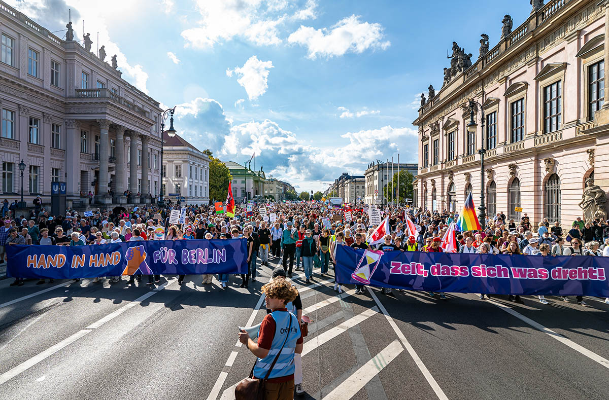 Farbfotografie einer großen Demonstration in Berlin. Tausende Menschen ziehen bei sonnigem Wetter über eine breite Straße zwischen prächtigen historischen Gebäuden. An der Spitze des Demonstrationszuges werden zwei große dunkelblaue Transparente getragen. Auf dem linken steht „Hand in Hand für Berlin“, auf dem rechten „Zeit, dass sich was dreht!“. Zahlreiche Demonstrierende tragen Plakate und bunte Fahnen, darunter Regenbogenflaggen. Im Vordergrund steht eine Person mit einer blauen Ordnerweste. Der Demonstrationszug erstreckt sich bis weit in den Hintergrund.