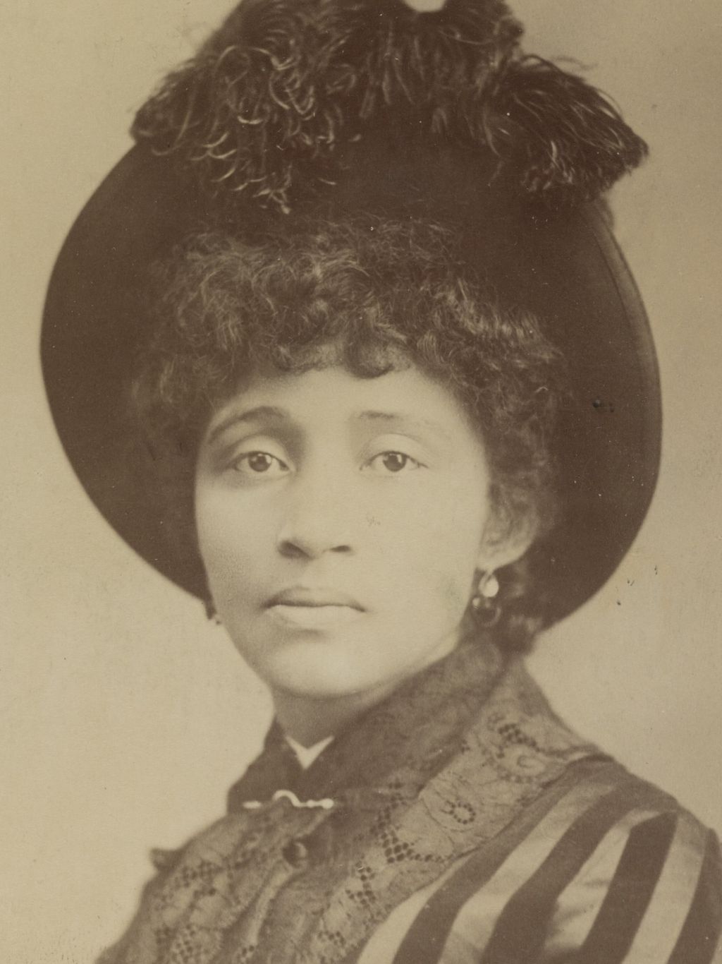 Historische, sepiafarbene Porträtfotografie der Anarchistin und Arbeiterrechtlerin Lucy Parsons aus dem Jahr 1886. Sie trägt einen großen, dunklen Hut mit üppigem Federschmuck und ein hochgeschlossenes, reich verziertes Kleid mit gestreiften Ärmeln. Ihr dunkles, lockiges Haar fällt bis an die Ohren, an denen kleine Ohrringe zu erkennen sind. Sie blickt mit ruhigem, ernstem Gesichtsausdruck direkt in die Kamera. Der helle, schlichte Hintergrund unterstreicht den Charakter der historischen Atelieraufnahme.