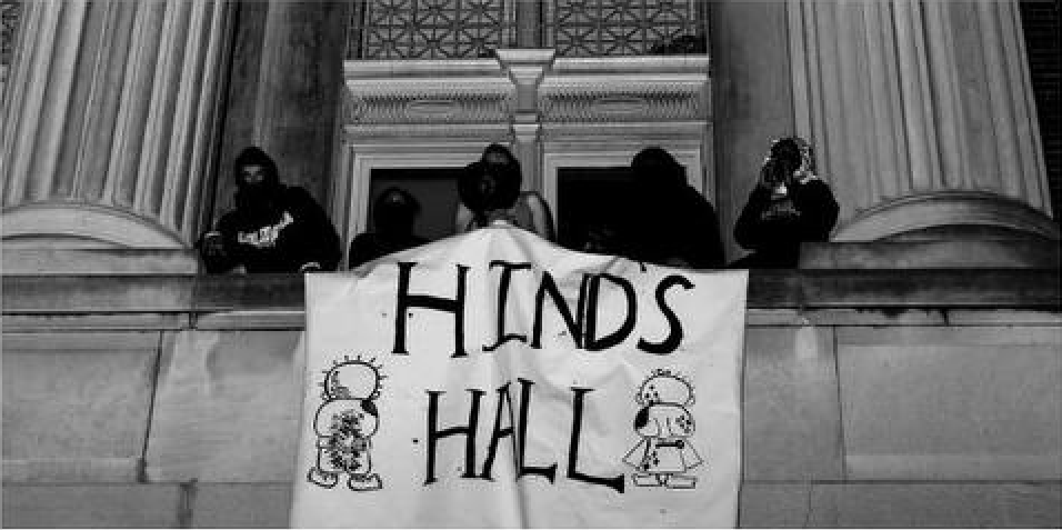 Das Foto zeigt zwei vermummte Personen, die ein Transparent mit dem Text "Hinds Hall" vom Balkon der Hamilton Hall hängen lassen