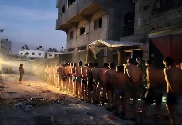 Das Foto zeigt eine lange Reihe mit dutzenden bis auf die Unterhode unbekleideter Manner, die von einer bewaffneten Person in Uniform bewacht werden.