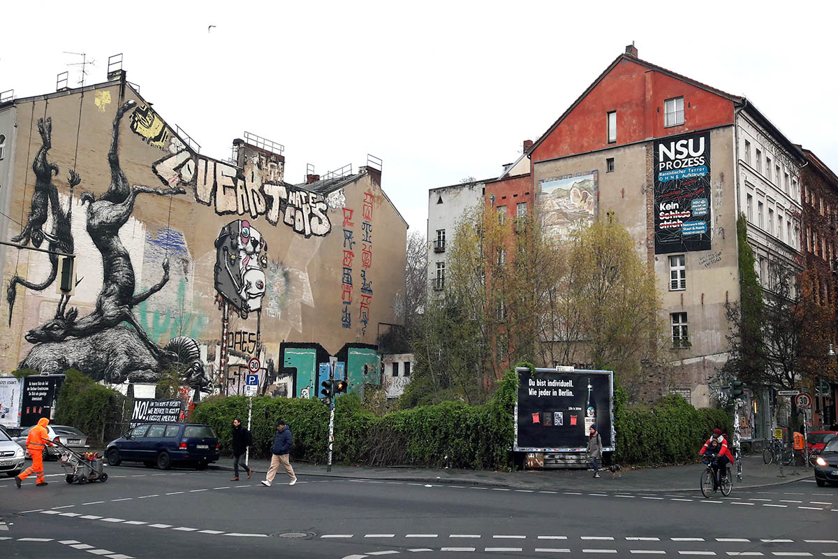 Das Foto zeigt die Brandmauern zweier Häuser. Auf der rechten Mauer hängt ein Plakat mit dem Text "NSU Prozess: Rassistischer Terror ohne Aufklärung. Kein Schlußstrich!". Auf der linken Brandmauer sind Grafitties zu sehen sowie große Gemälde mit an den Hinterbeinen aufgehängten Tieren
